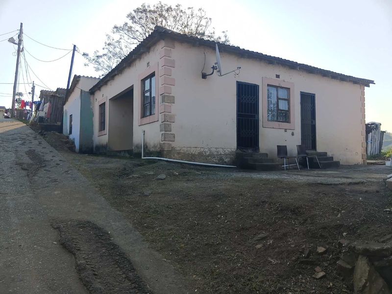 Single-story residential house featuring a tiled roof, security gates, and exterior plaster finish, situated on an unpaved access road.