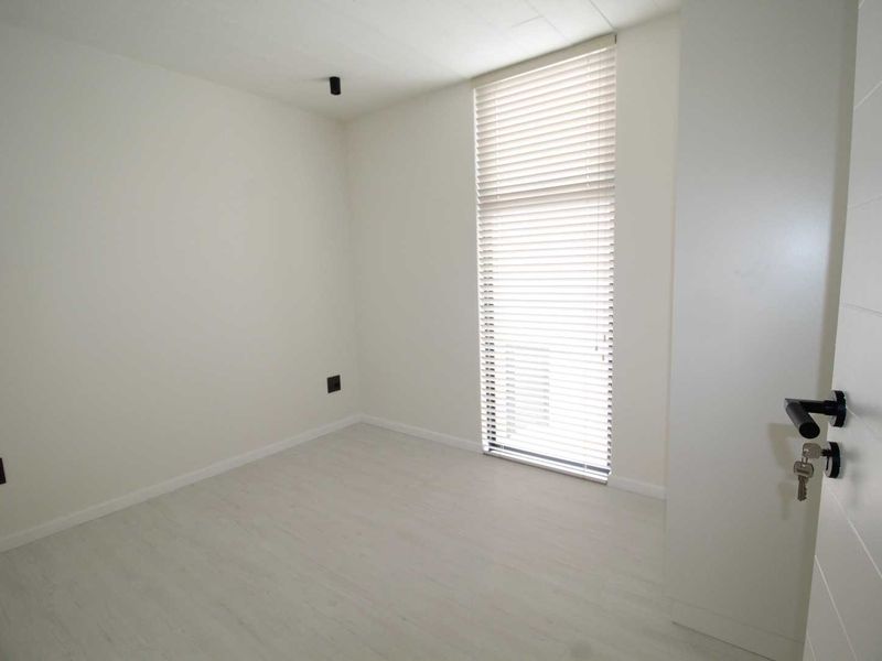 A bright, modern, and empty bedroom featuring light laminate flooring, neutral walls, and fitted venetian blinds.