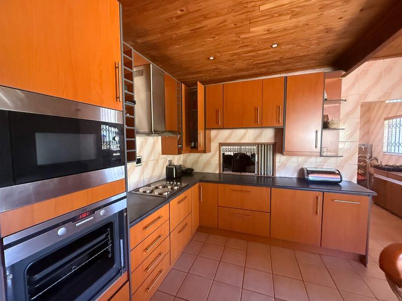 A functional kitchen featuring warm-toned wooden cabinetry, a timber-clad ceiling, tiled flooring, and integrated stainless steel appliances including an oven and hob.