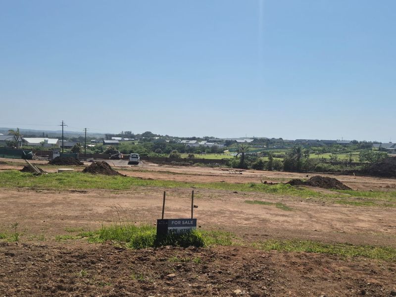 A wide-angle view of a vacant residential plot of land with a for sale sign, featuring surrounding neighborhood views and clear sky.