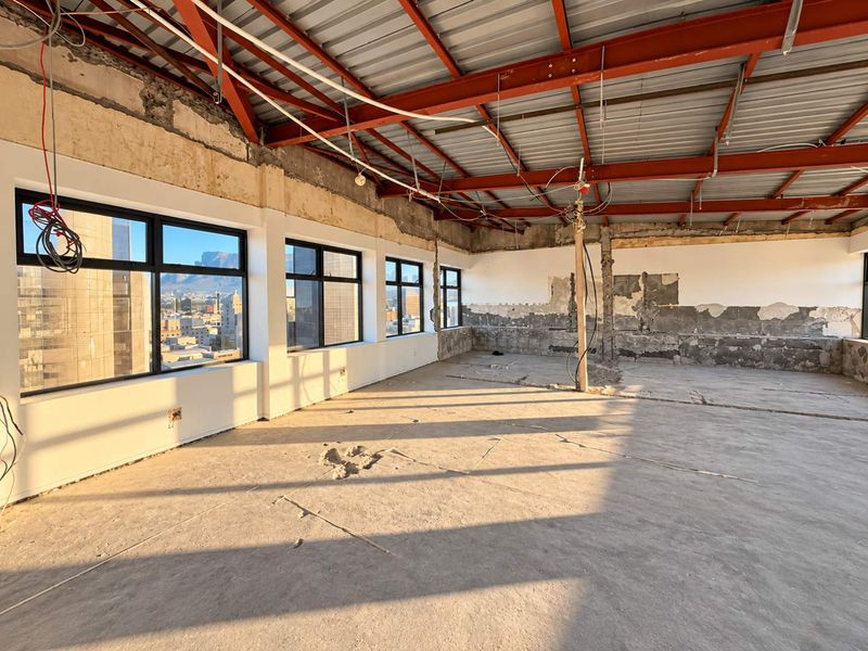 A spacious, open-plan commercial interior currently under renovation, featuring exposed steel roof trusses, concrete floors, and large windows with city views.