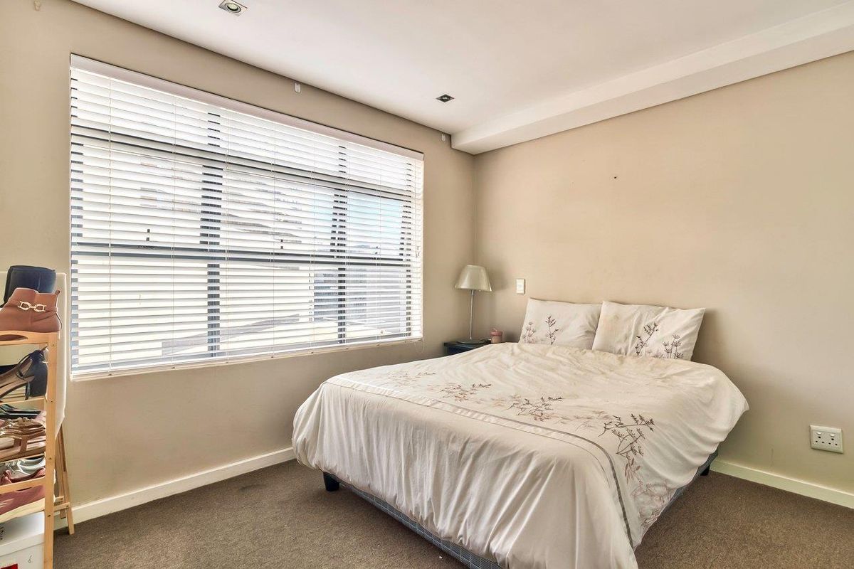 A well-lit bedroom featuring neutral wall tones, carpeted flooring, and large windows fitted with venetian blinds.