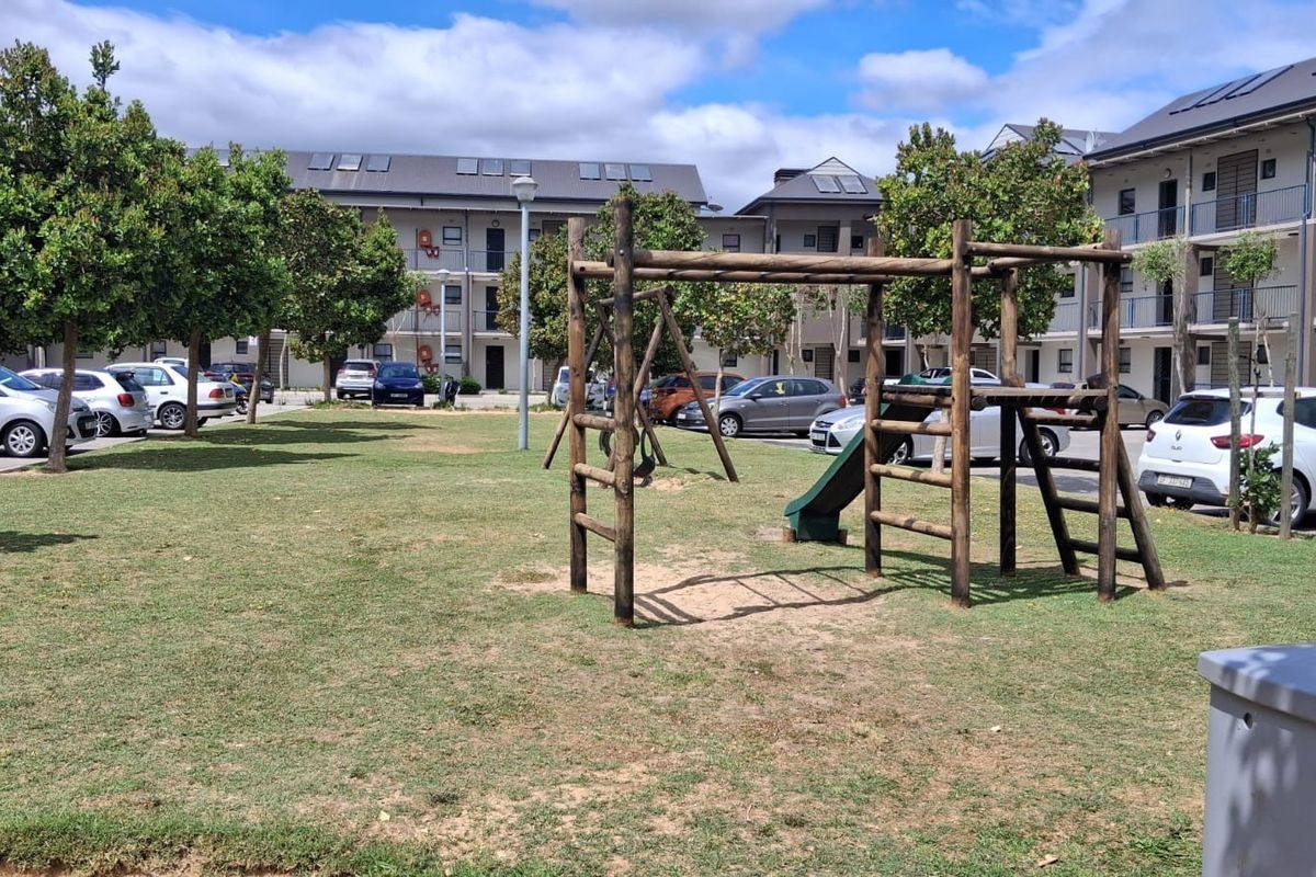 Communal garden area featuring a wooden playground structure, landscaped trees, and adjacent paved parking for the apartment complex.
