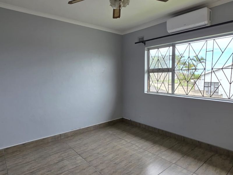 A clean, empty bedroom featuring tiled flooring, a ceiling fan, and a wall-mounted air conditioning unit, with security bars installed on the window.