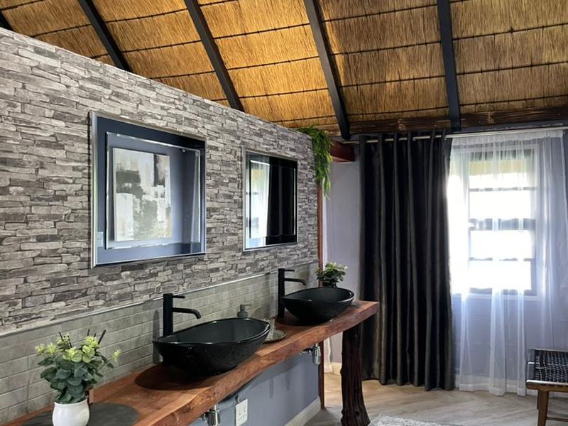 A stylish bathroom featuring a double vanity with vessel sinks, stone veneer wall accents, and a high vaulted thatched ceiling.