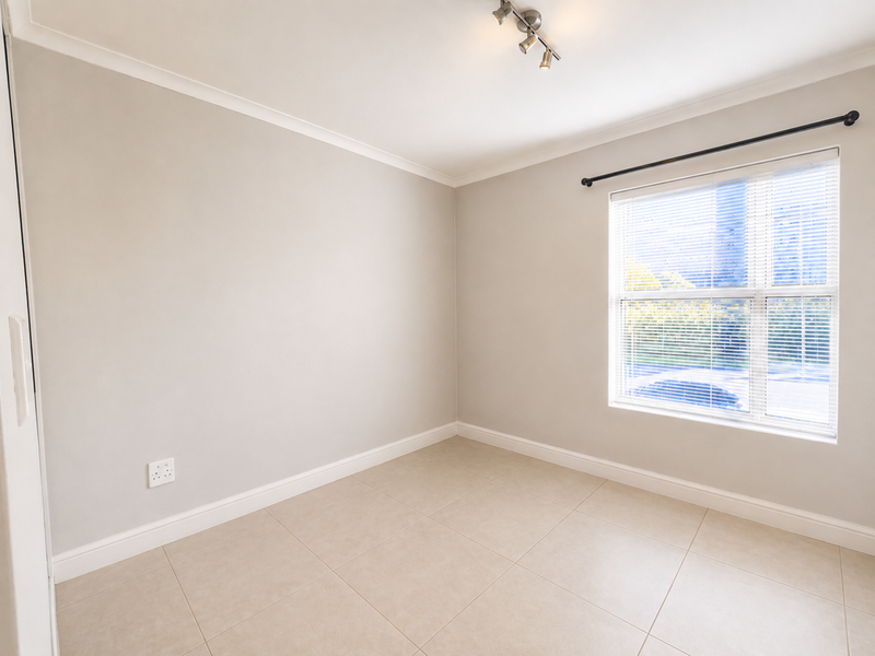 A bright, empty bedroom featuring neutral tiled flooring, clean walls, and a window fitted with venetian blinds and a curtain rod.