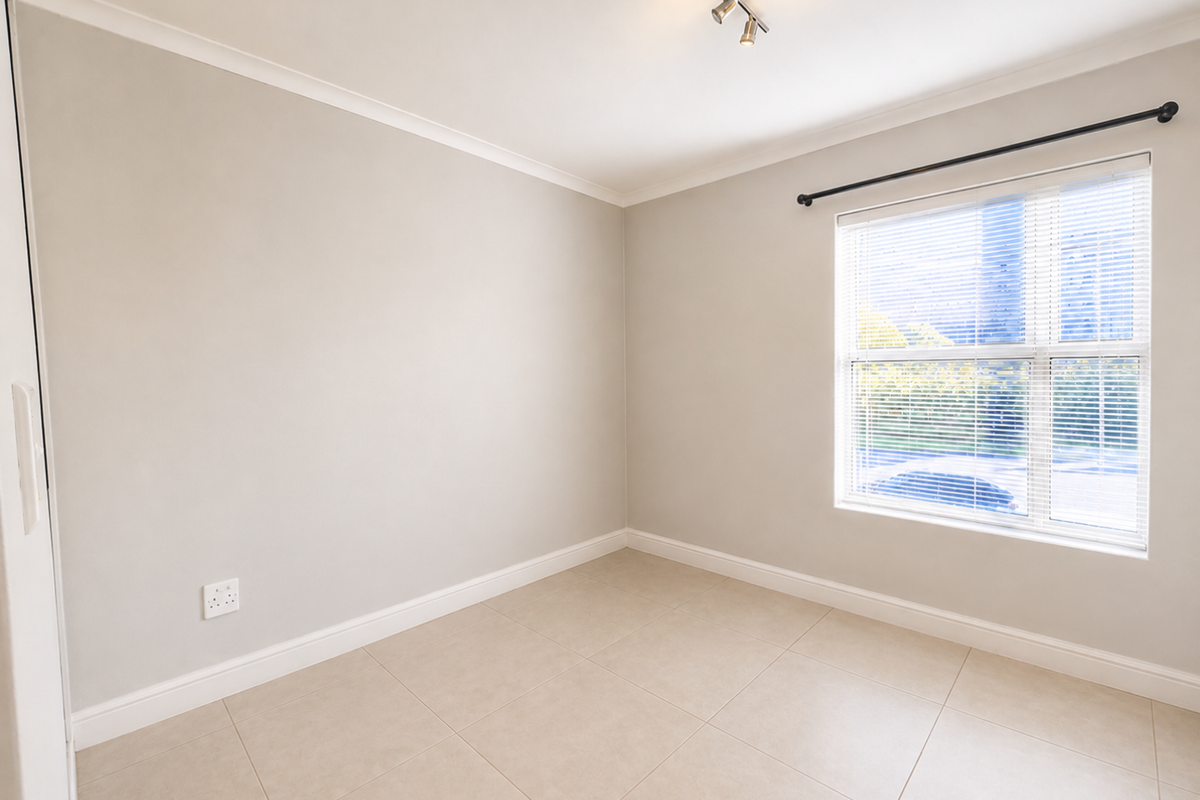 A bright, empty bedroom featuring neutral tiled flooring, clean walls, and a window fitted with venetian blinds and a curtain rod.