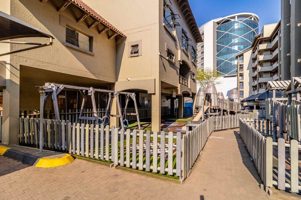 A well-maintained outdoor communal area featuring a paved walkway, fenced playground equipment, and artificial turf situated between apartment building blocks.