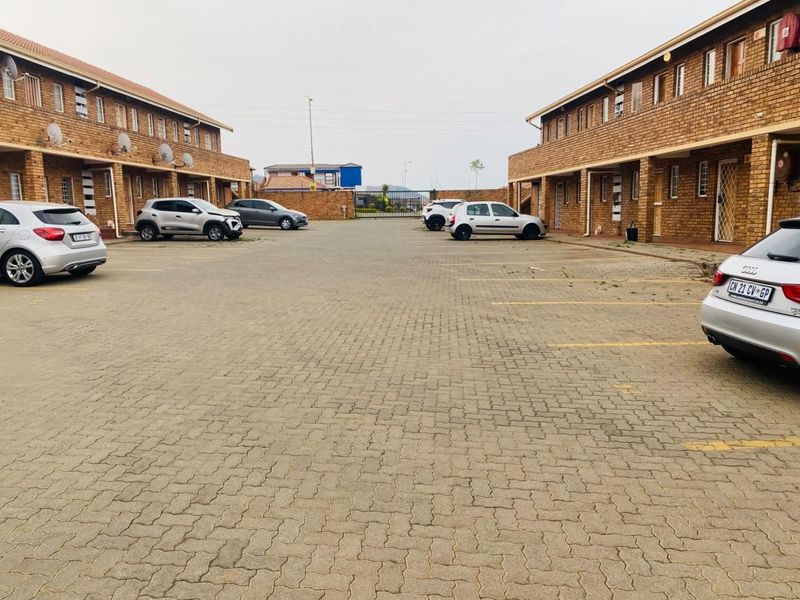 Exterior view of an apartment complex featuring a central paved parking area between two brick-faced residential buildings.