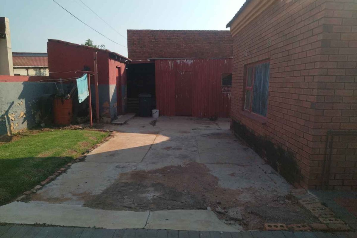 Rear exterior view showing a concrete courtyard, brick house facade, and secondary metal outbuilding structure with signs of significant wear and weathering.