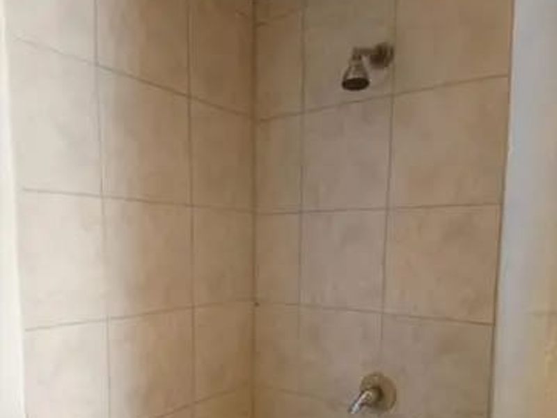 A basic shower enclosure featuring neutral ceramic wall tiles, a standard shower head, and a wall-mounted mixer tap.