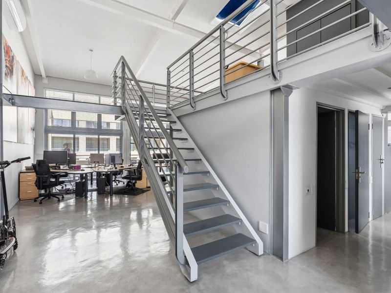 Modern open-plan office space featuring a mezzanine level, polished concrete floors, exposed structural steel elements, and a central staircase.