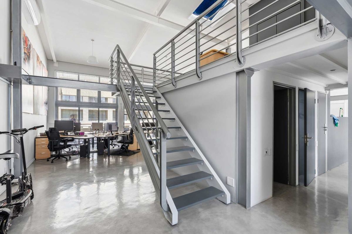 Modern open-plan office space featuring a mezzanine level, polished concrete floors, exposed structural steel elements, and a central staircase.