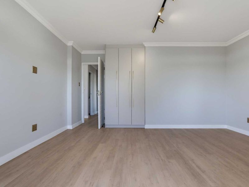 A bright, empty bedroom featuring modern laminate flooring, built-in wardrobes, and contemporary track lighting, finished with clean crown molding.