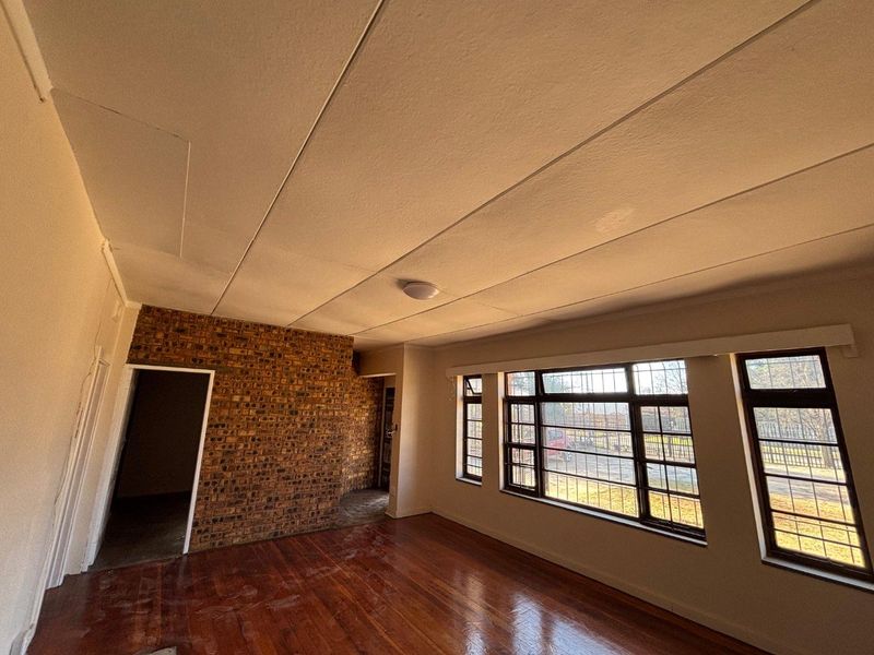 An empty living room featuring an exposed brick wall, hardwood flooring, and large steel-framed windows with security bars.