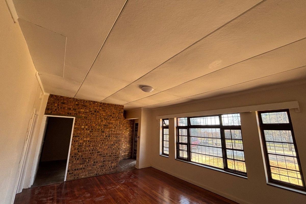 An empty living room featuring an exposed brick wall, hardwood flooring, and large steel-framed windows with security bars.