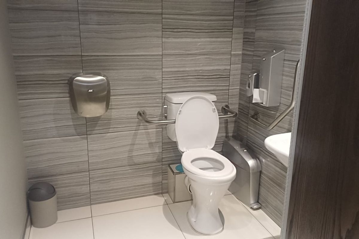 A clean, modern accessible bathroom featuring tiled walls, a toilet with grab rails, and wall-mounted utility fixtures.