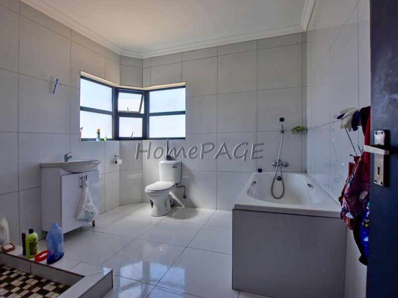 A bright, tiled bathroom featuring a corner window, a vanity basin, a standard toilet, and a built-in bathtub with a shower mixer.
