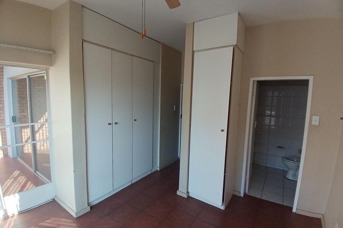 A spacious bedroom featuring built-in cupboards, tiled flooring, a ceiling fan, and direct access to an en-suite bathroom and a private balcony.