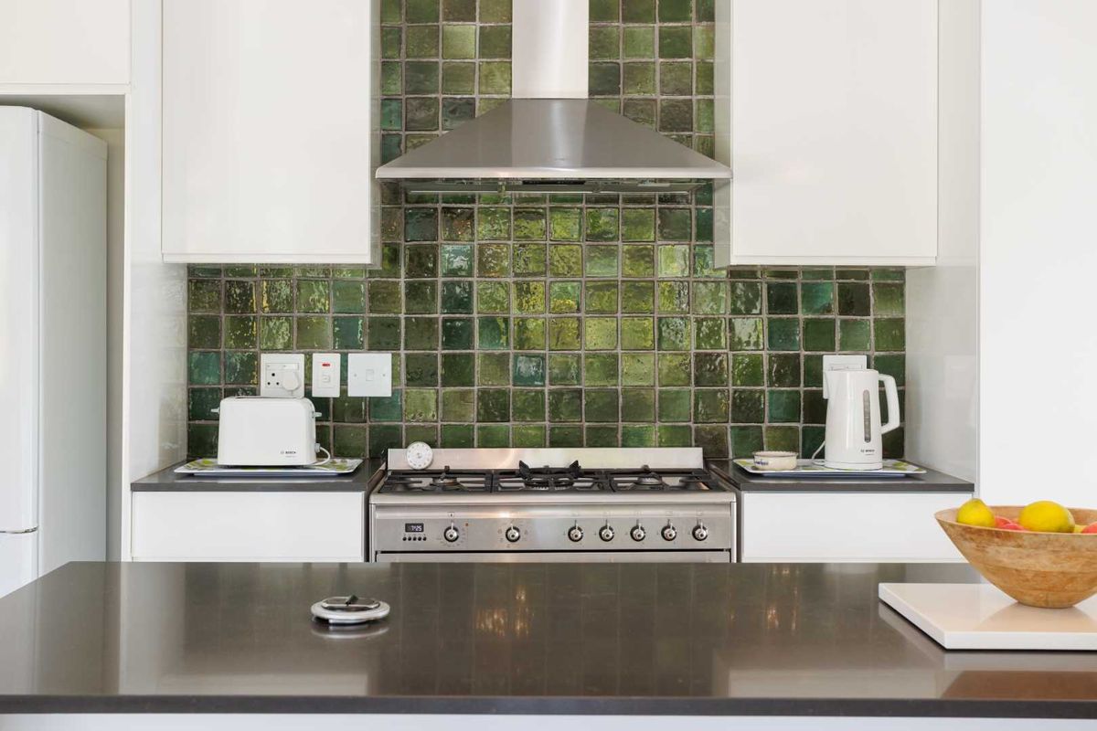 Modern kitchen featuring a stainless steel gas range, extractor fan, and a striking green tiled backsplash with white cabinetry and dark countertops.