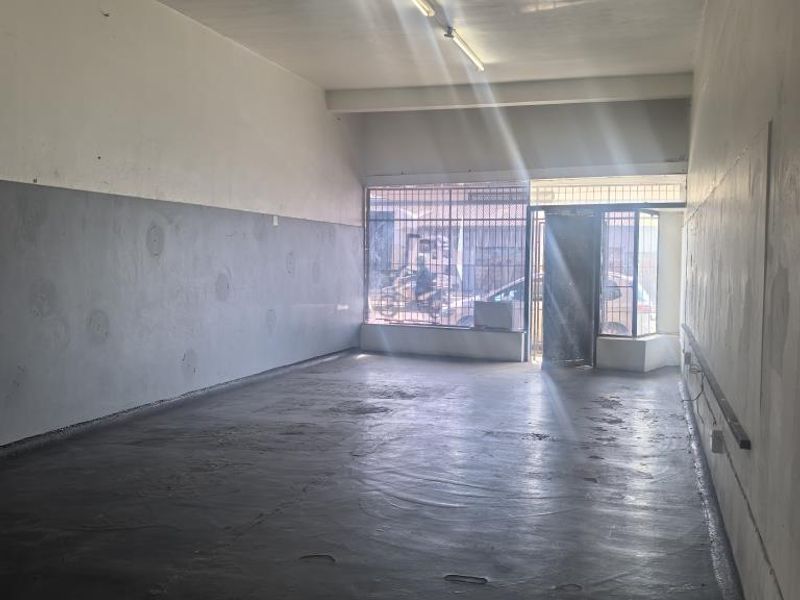 An empty, open-plan commercial space featuring concrete flooring, fluorescent lighting, and secure street-facing access with security bars.