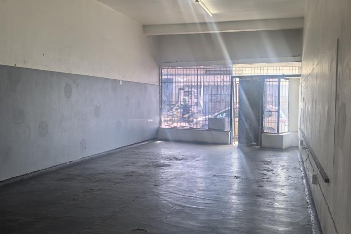 An empty, open-plan commercial space featuring concrete flooring, fluorescent lighting, and secure street-facing access with security bars.