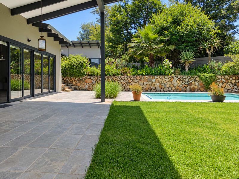 A well-maintained outdoor entertainment area featuring a tiled patio, swimming pool, and a stone retaining wall set against a lush garden.