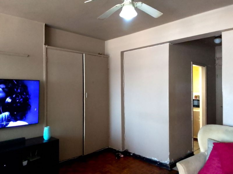 Living room featuring parquet flooring, a ceiling fan, and built-in storage cupboards, with visible wear to the skirting boards and wall finishes.