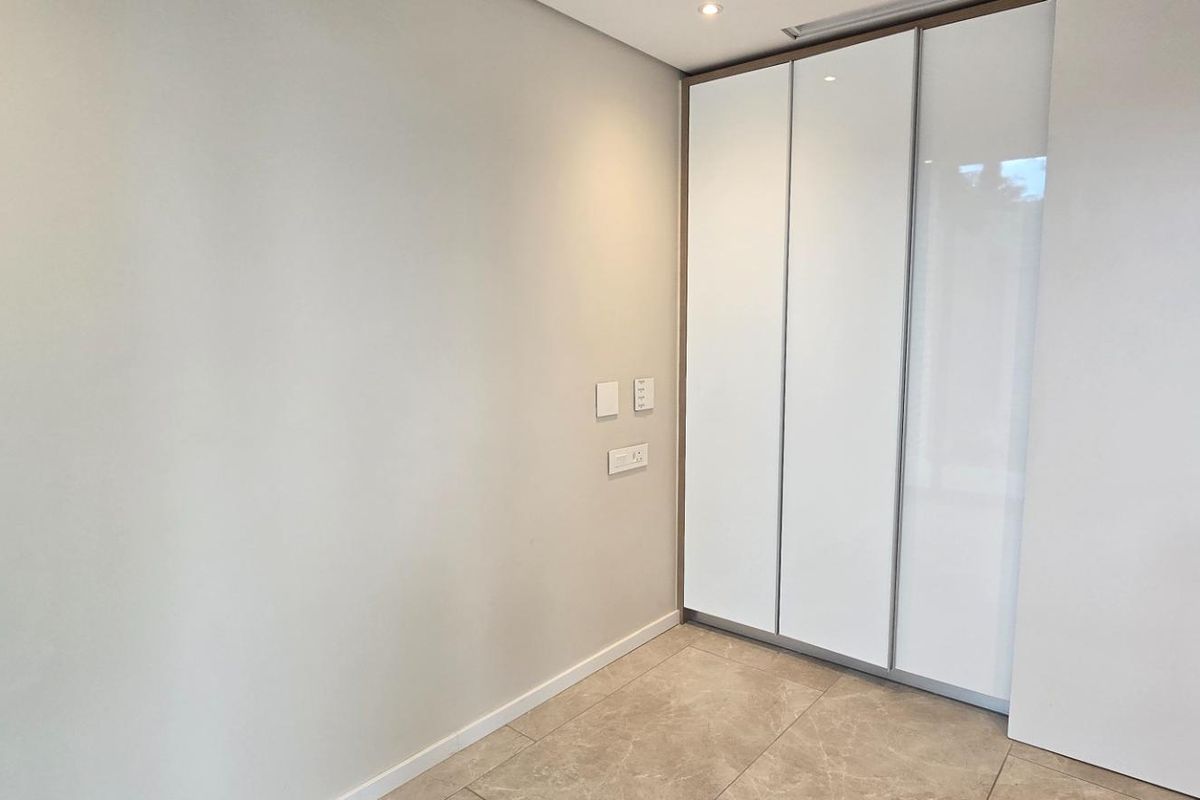 A clean, modern bedroom interior featuring high-gloss built-in wardrobes, neutral tiled flooring, and recessed ceiling lighting.