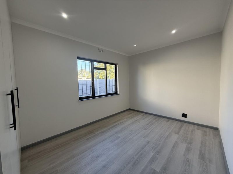 A bright, empty bedroom featuring modern laminate flooring, recessed downlighting, and a built-in wardrobe with dark hardware.
