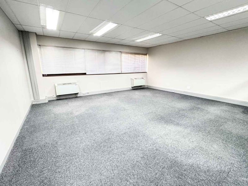 A vacant, open-plan office space featuring neutral carpeting, a suspended ceiling with recessed lighting, and wall-mounted HVAC units for climate control.