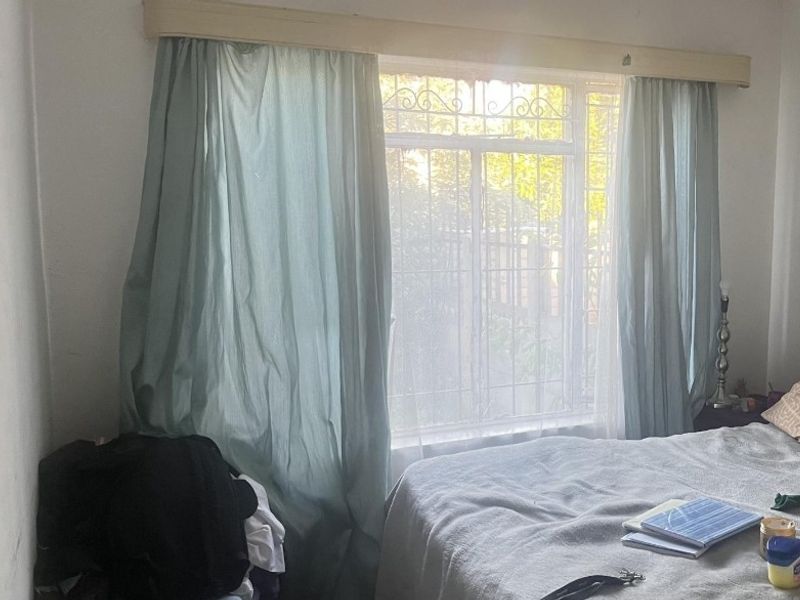 A bedroom featuring a window with security burglar bars and a wooden curtain pelmet, providing natural light and exterior views.