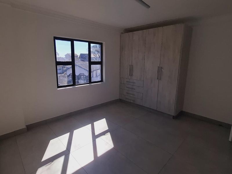 A bright, empty bedroom featuring neutral tiled flooring, built-in wooden cupboards, and a window providing natural light.