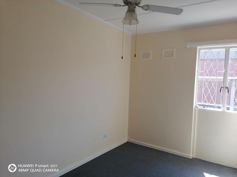 A clean, empty bedroom featuring neutral walls, grey carpeting, a ceiling fan, and a window fitted with security bars.