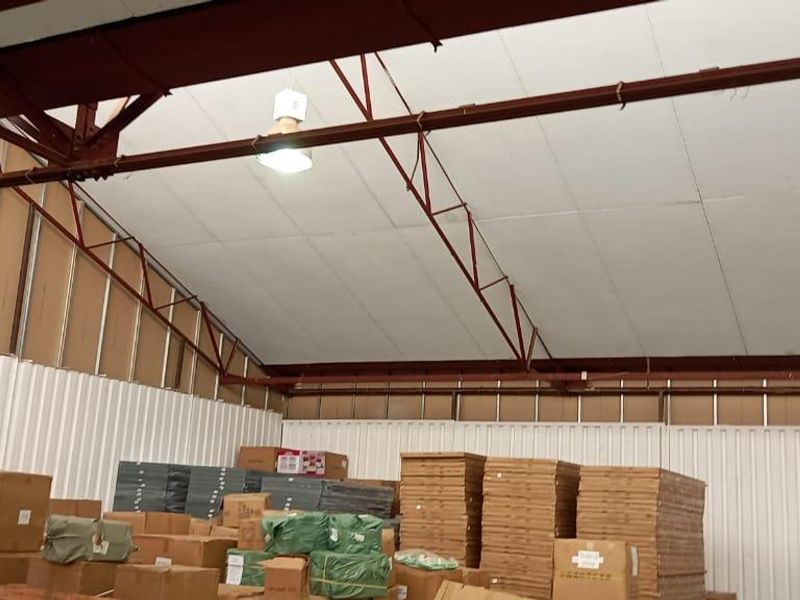 Spacious industrial warehouse interior featuring high ceilings with exposed steel trusses, corrugated metal walls, and functional overhead industrial lighting.