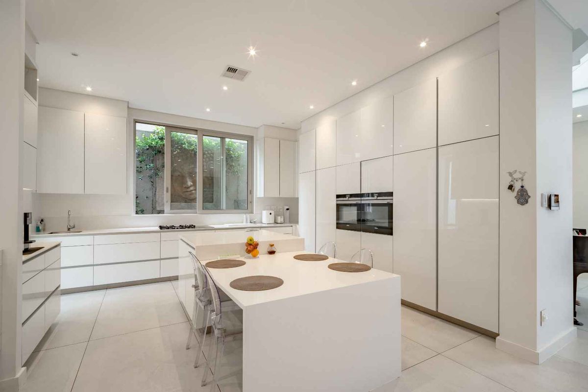 Modern, minimalist kitchen featuring high-gloss white cabinetry, a central island with integrated seating, built-in oven, and large windows overlooking a private garden.