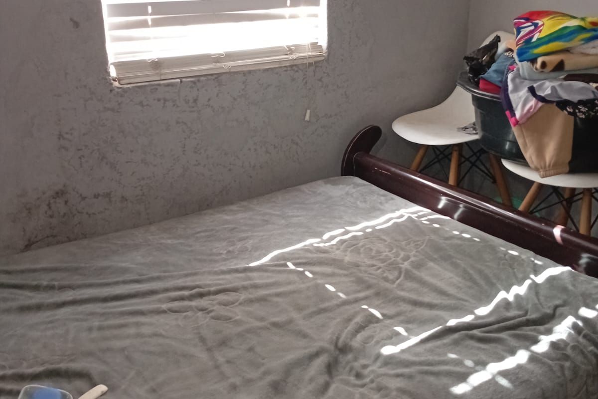 A bedroom featuring a window with venetian blinds and visible wall surface damage requiring maintenance.