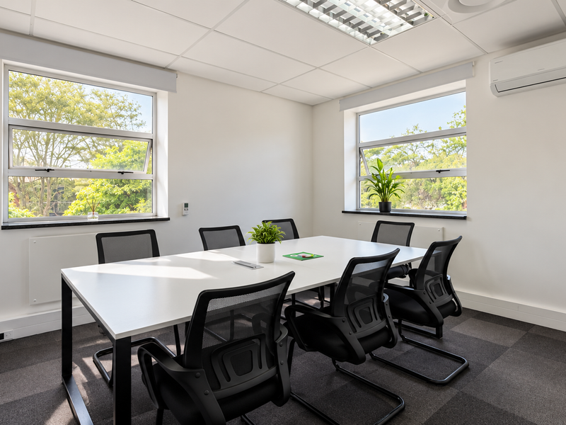 A bright, professional office meeting room featuring carpeted flooring, suspended ceiling with recessed lighting, split-system air conditioning, and large windows providing natural light.