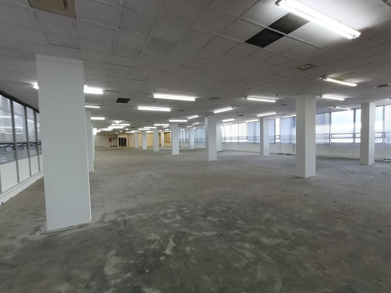 A large, open-plan office space featuring structural columns, suspended ceilings with fluorescent lighting, and expansive windows with vertical blinds.