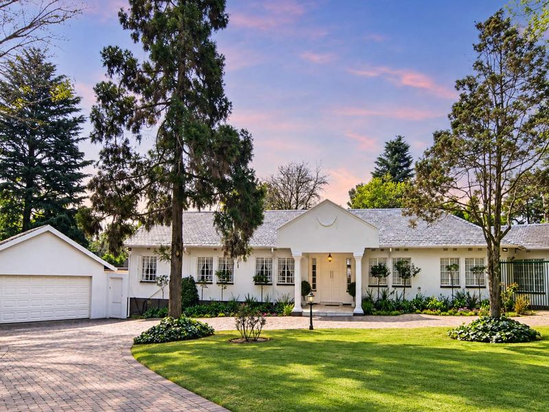 A beautifully maintained single-story residence featuring a classic white facade, covered entrance portico, paved driveway, and a detached garage set within a lush garden.