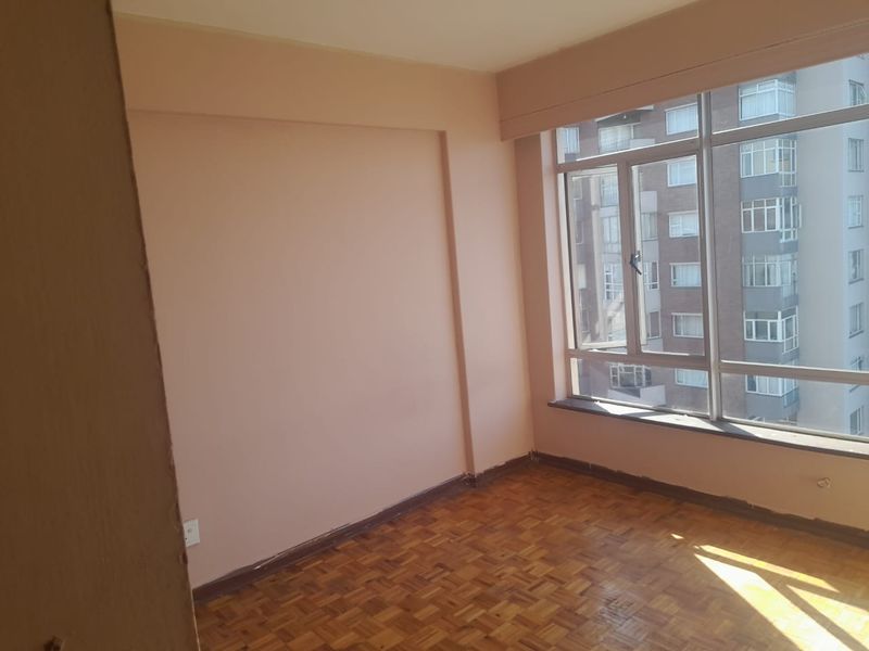 An empty bedroom featuring classic parquet flooring and a large window providing natural light and an urban outlook.