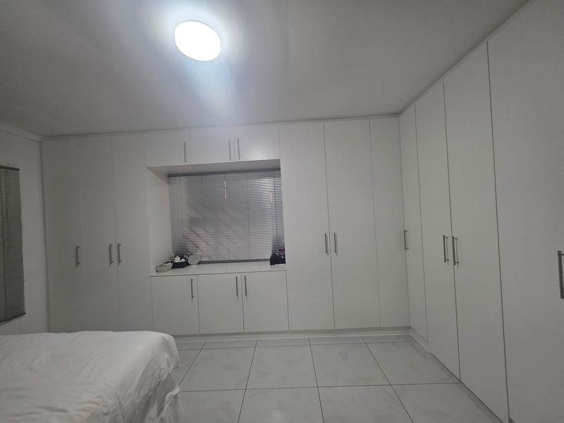 A spacious bedroom featuring extensive white built-in cupboards, tiled flooring, and a window with venetian blinds.