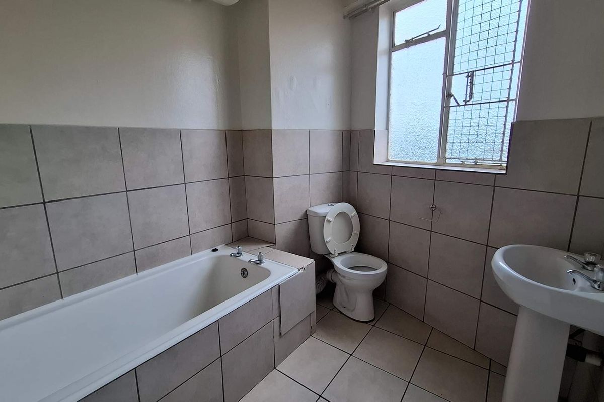 A functional bathroom featuring a tiled bathtub, pedestal basin, and toilet, with neutral wall and floor tiling and a security-grilled window.