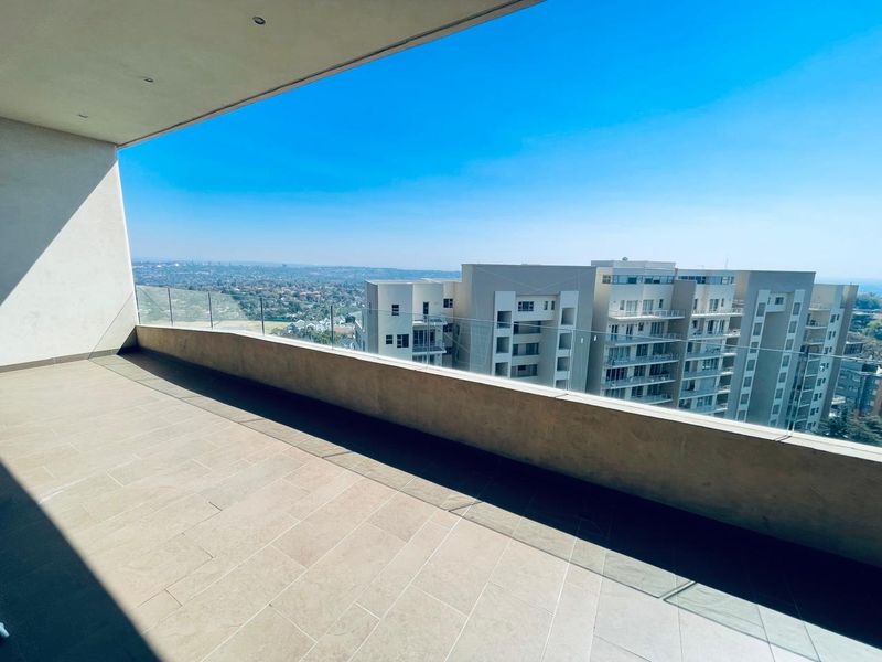 A spacious, modern balcony featuring a sleek glass balustrade, tiled flooring, and expansive elevated views of the surrounding cityscape.