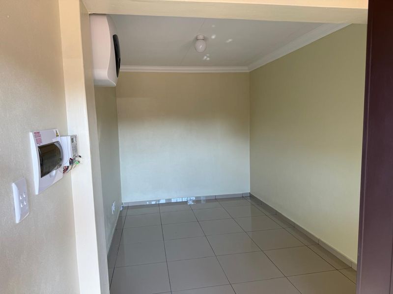 An empty, neutral-toned living area featuring tiled flooring, a wall-mounted heater, and a visible electrical distribution board.