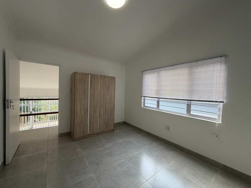 A clean, neutral bedroom featuring large format floor tiles, a built-in wardrobe, and a window fitted with venetian blinds and security bars.