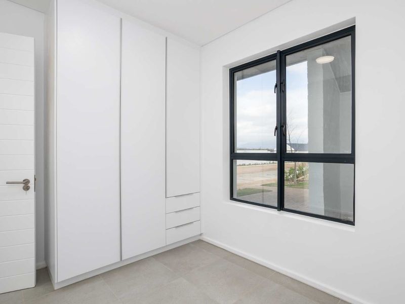 A bright, modern bedroom featuring clean white built-in cupboards, neutral tiled flooring, and a large window with a view.