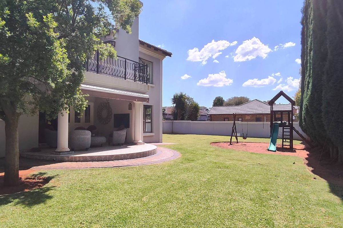 Spacious rear garden featuring a covered patio with a balcony above, paved flooring, and a well-maintained lawn enclosed by a perimeter wall.