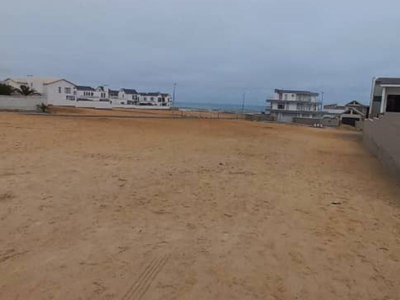 A vacant residential plot of land featuring sandy terrain, offering a clear view of the surrounding neighborhood and distant coastal horizon.