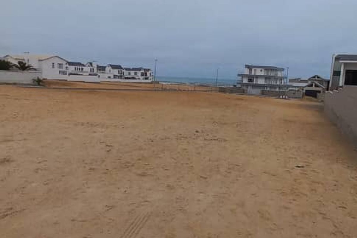 A vacant residential plot of land featuring sandy terrain, offering a clear view of the surrounding neighborhood and distant coastal horizon.